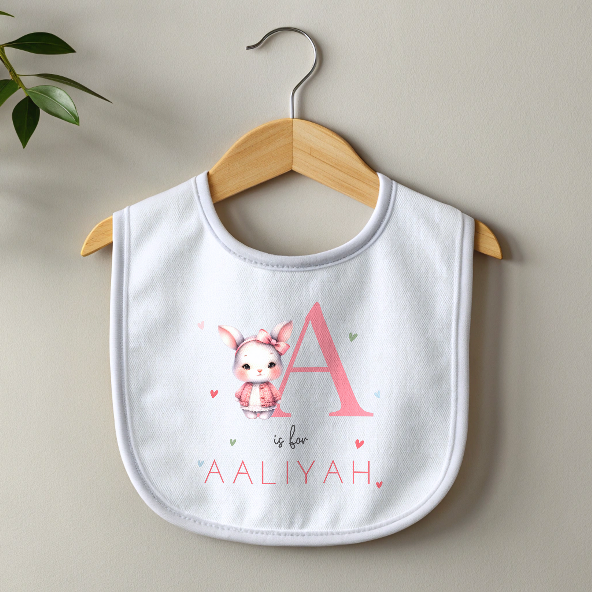 personalised baby outfit: An adorable baby girl wearing a pink sleepsuit for, featuring a cute dusky pink rabbit design. This personalised outfit is available as a sleepsuit, bodysuit, T-shirt, or bib, making it a perfect new baby gift. Ideal for coming home outfits or gender reveal announcements. It has the baby&#39;s initial and name along with the pink rabbit design.