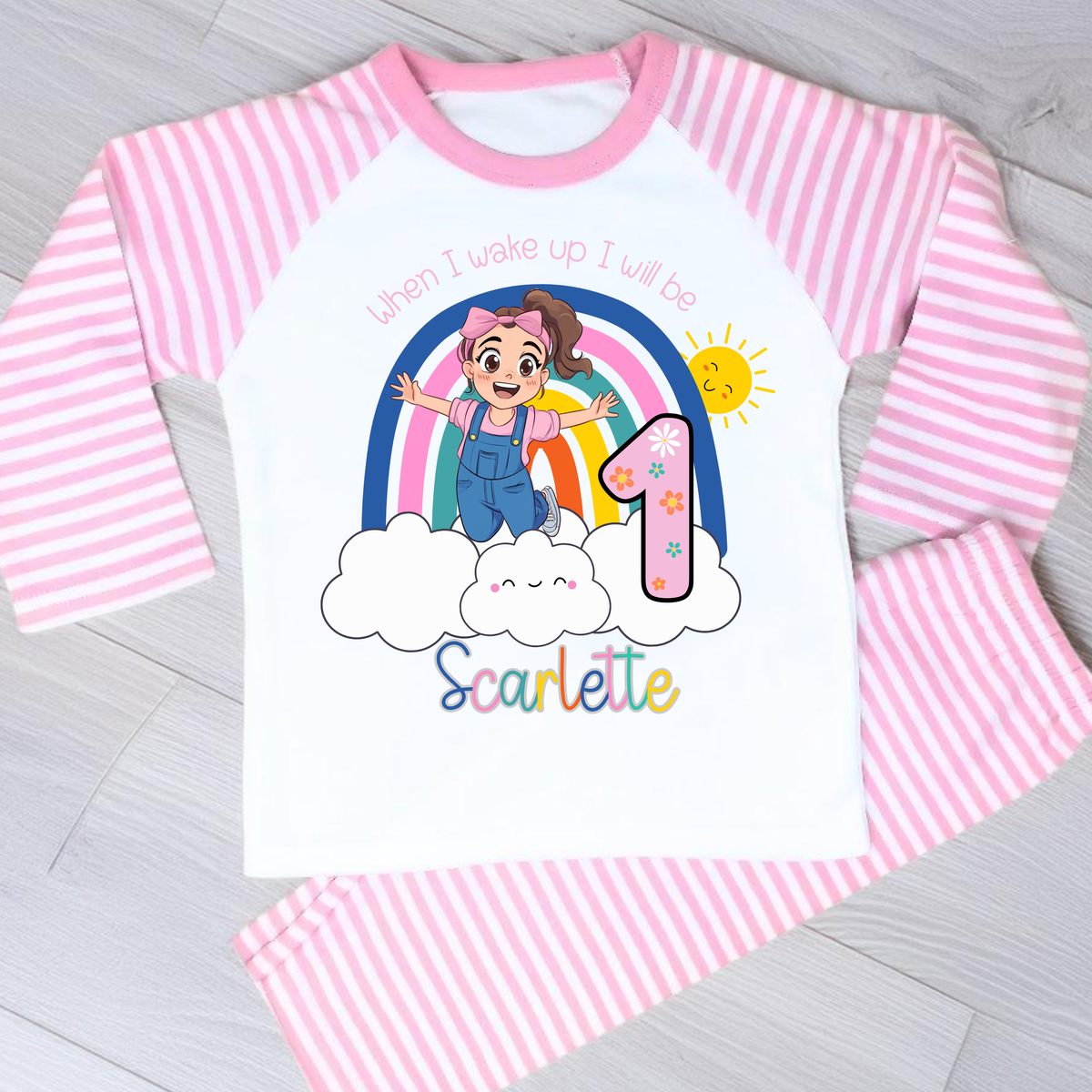 Personalised Ms Rachel Birthday PJs for Girls, When I Wake up Ms Rachel Pyjamas AishaBaileyDesigns: A children's pajama set laid flat on a light gray wood surface, featuring a white long-sleeve top with pink and white vertical stripes, a pink collar, and a colorful first birthday graphic on the front with the text 'When I wake up I will be 1 Scarlett' and a cartoon girl under a rainbow. Matching pink-and-white striped pajama pants are visible.