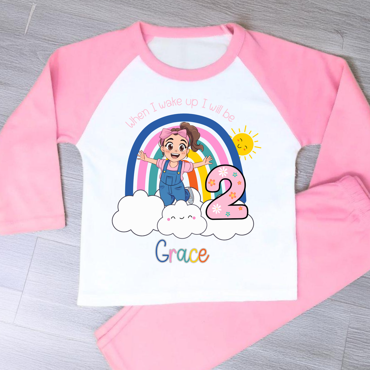 Personalised Ms Rachel Birthday PJs for Girls, When I Wake up Ms Rachel Pyjamas AishaBaileyDesigns: A pink and white raglan long-sleeve pajama set laid flat on a gray wood floor. The white front panel features a colorful cartoon graphic of a girl in blue overalls under a rainbow and sun, with the text 'when I wake up I will be 2 Grace'.