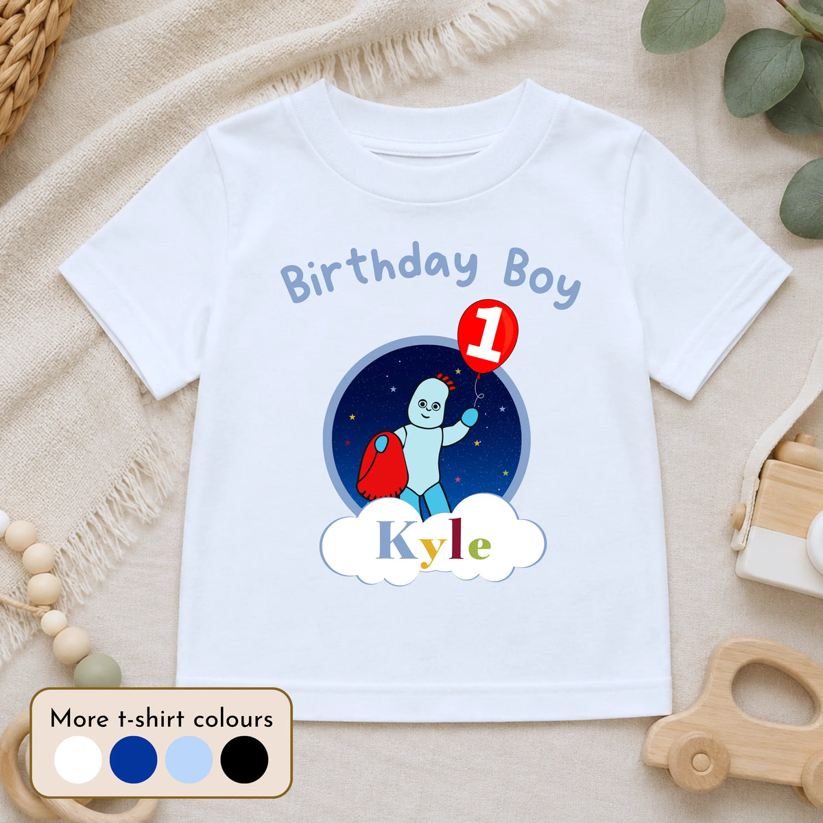 A personalised white short-sleeve baby t-shirt with a 'Birthday Boy Kyle' first birthday graphic, displayed on a neutral beige fabric background with wooden beads and a toy car. In the Night Garden theme with Iggle Piggle.