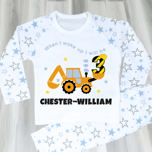 A flat lay blue Pyjama set with a yellow excavator design. The excavator has a black bucket and a black age number on the side. The text 'When I Wake up I will be' is above the excavator and the child's name is below the excavator Digger Construction. Boys personalised birthday gift, Tractor birthday pyjamas, boys custom pyjamas with name, When I wake up construction digger, birthday pyjamas with name