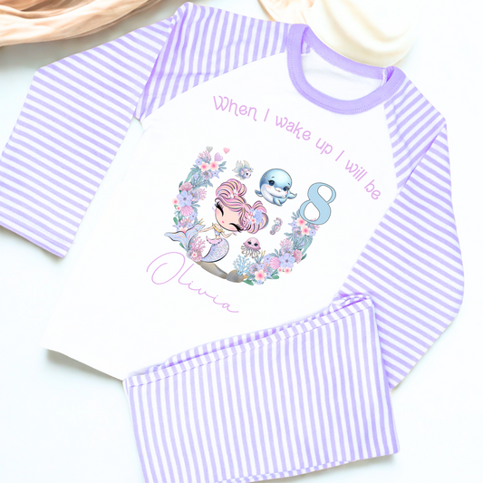 Mermaid birthday pyjamas, personalised for girls: A pair of mermaid birthday pyjamas in lilac and pink, featuring a glitter-like design. The top displays the phrase “When I Wake Up I Will be,” personalized with a girl's name and age. Ideal for young mermaid enthusiasts, these Pyjamas make a magical birthday gift for girls, when i wake up mermaid