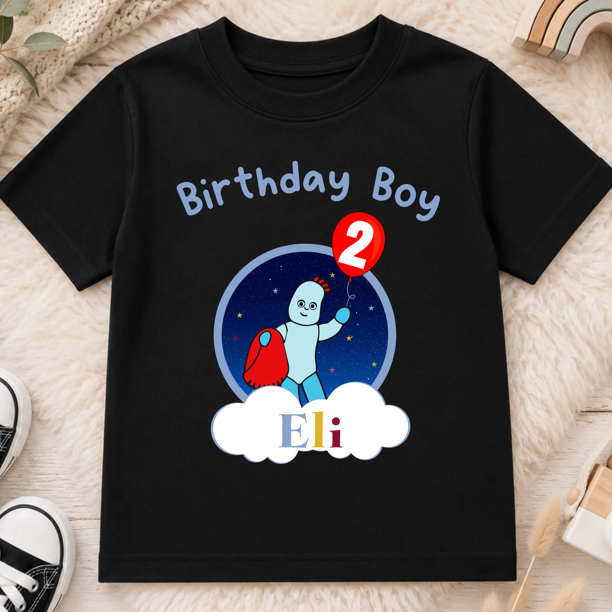 A personalised black short-sleeve baby t-shirt with a 'Birthday Boy Eli' second birthday graphic, displayed on a neutral beige fabric background with wooden beads and a toy car. In the Night Garden theme with Iggle Piggle.
