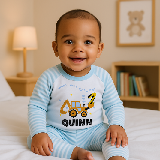 A baby boy wearing blue Pyjama set with a yellow excavator design. The excavator has a black bucket and a black age number on the side. The text 'When I Wake up I will be' is above the excavator and the child's name is below the excavator Digger Construction. Boys personalised birthday gift, Tractor birthday pyjamas, boys custom pyjamas with name, When I wake up construction digger, birthday pyjamas with name